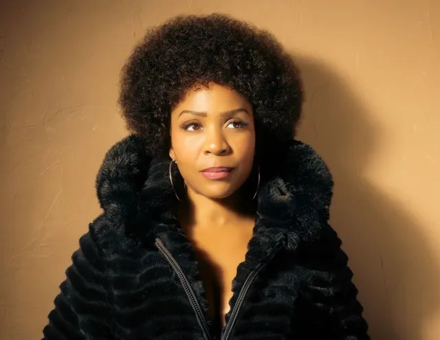 Divine Brown as Minnie Riperton in a black faux-fur coat