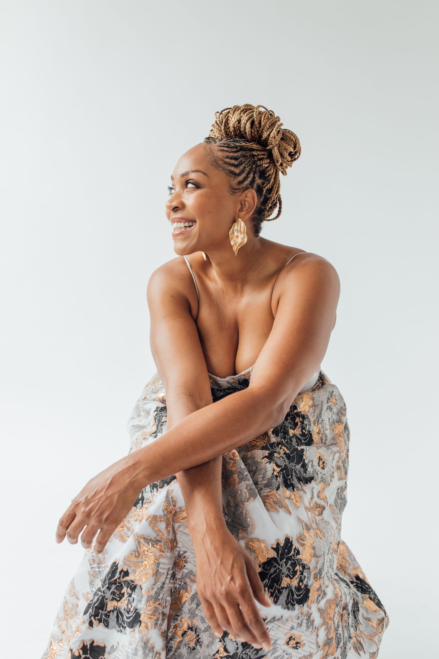 Divine Brown crouched in a white blazer and gold heels